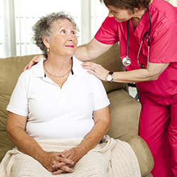 Woman with stethoscope around neck and hands on shoulders of mature woman sitting with blanket on lap