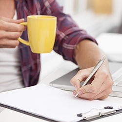 Person writing on a clipboard next to a laptop on a desk
